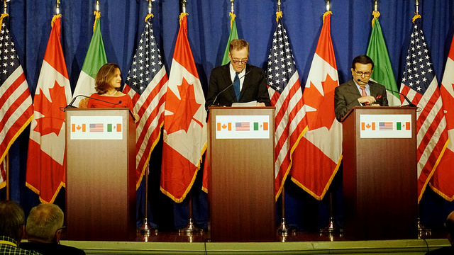 Three people stand at podiums with flags behind them